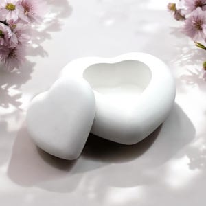 Op de afbeelding: Een witte keramische hartvormige doos met een deksel. De doos is open en het deksel ligt ernaast. De doos staat op een wit oppervlak.