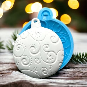 Può includere: Uno stampo in silicone bianco per realizzare decorazioni natalizie. Lo stampo ha la forma di un ornamento rotondo con un design a spirale. Lo stampo è mostrato con un ornamento bianco che è stato realizzato usando lo stampo.