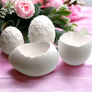 Könnte beinhalten: Drei weiße, eiförmige Behälter aus Keramik. Zwei der Behälter sind ganz, einer ist zerbrochen und zeigt das Innere. Die Behälter stehen auf einer rosa Oberfläche.