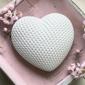 Pode incluir: Um objeto branco em forma de coração com uma superfície texturizada, repousando sobre uma bandeja rosa. Delicadas flores cor-de-rosa estão espalhadas ao redor do coração, criando uma estética suave e romântica. O coração tem um padrão de covinhas.