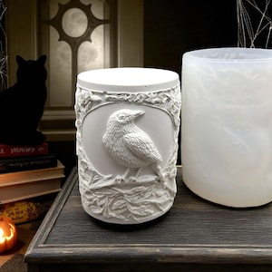 May include: A white cylindrical candle holder featuring a detailed relief of a bird on a branch, framed by leaves. Beside it is a translucent white container. The scene has a Halloween theme.