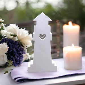 Puede incluir: Figurita de faro de madera blanca con un recorte en forma de corazón, sentada sobre un camino de mesa morado junto a dos velas blancas.