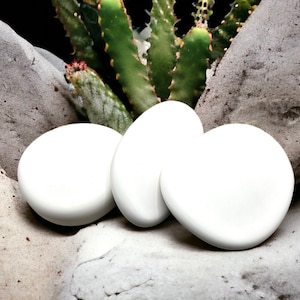 May include: Three smooth, white, oval-shaped stones arranged on a light grey, textured surface. The stones are smooth and appear to be made of a polished material. The background features a green cactus plant.