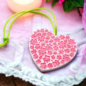 Op de afbeelding: Een roze keramische hartvormige hanger met een groene touwtje om op te hangen. Het hart is bedekt met een patroon van roze poot afdrukken.