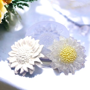 May include: Two white resin sunflower ornaments, one with a yellow center and the other with a white center, next to a silicone mold of a sunflower.