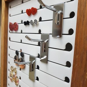 May include: White pegboard with three silver metal hooks for hanging earrings. The pegboard has black dots and is framed with a brown wood frame.  A variety of earrings are hanging on the hooks.