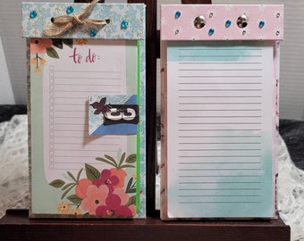 May include: Two floral patterned notepad holders with to-do lists. The left holder has a green and blue floral pattern with a to-do list and a clip. The right holder has a pink and blue floral pattern with a blank notepad.