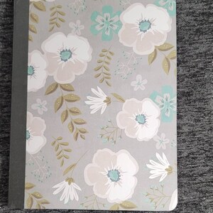 May include: A gray and white notebook with a floral pattern. The flowers are white with a light blue center and are surrounded by green leaves. The notebook has a black spine.