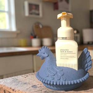 Puede incluir: Un jabonero de cer&aacute;mica azul con forma de gallina con un frasco de jab&oacute;n de manos espumoso de leche y miel de pistacho. El frasco de jab&oacute;n es blanco con una bomba beige. El soporte para gallinas est&aacute; sobre una superficie de madera.