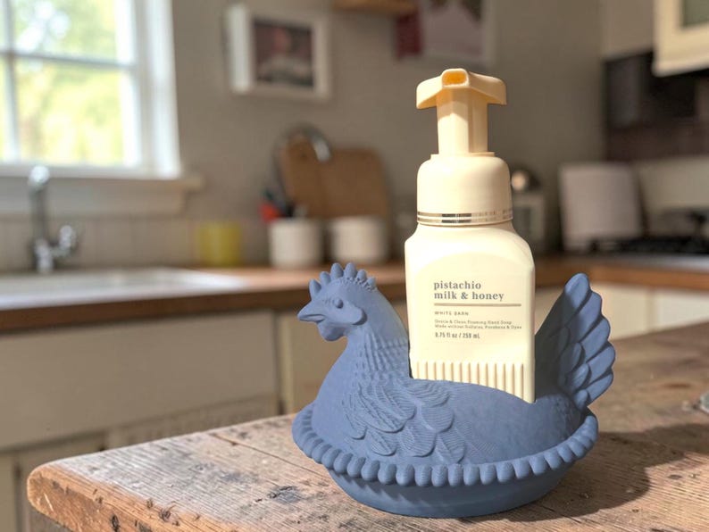 Puede incluir: Un jabonero de cer&aacute;mica azul con forma de gallina con un frasco de jab&oacute;n de manos espumoso de leche y miel de pistacho. El frasco de jab&oacute;n es blanco con una bomba beige. El soporte para gallinas est&aacute; sobre una superficie de madera.