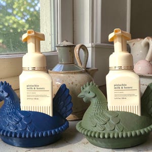 Puede incluir: Dos dispensadores de jab&oacute;n decorativos con forma de gallina, uno azul y otro verde, cada uno con una botella de jab&oacute;n de manos "pistacho, leche y miel". Los dispensadores est&aacute;n en un alf&eacute;izar, con otros objetos decorativos en el fondo.