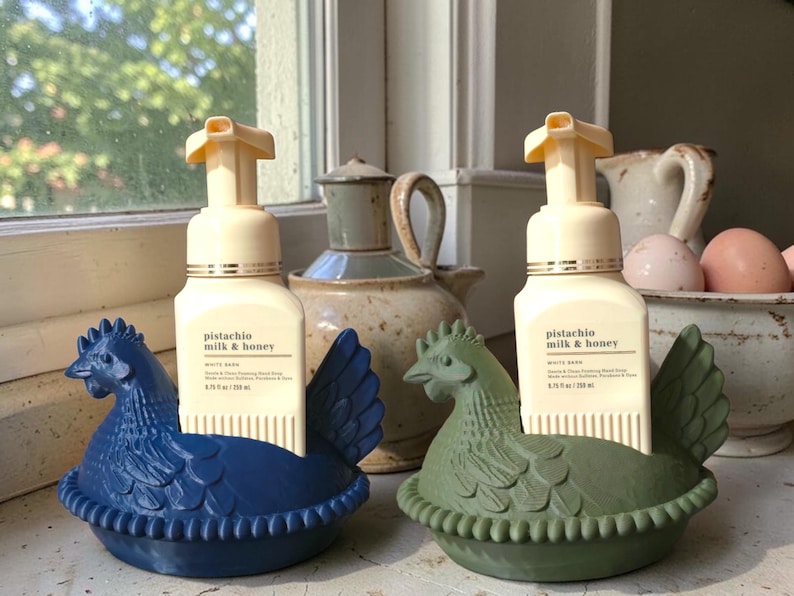 Puede incluir: Dos dispensadores de jab&oacute;n decorativos con forma de gallina, uno azul y otro verde, cada uno con una botella de jab&oacute;n de manos "pistacho, leche y miel". Los dispensadores est&aacute;n en un alf&eacute;izar, con otros objetos decorativos en el fondo.