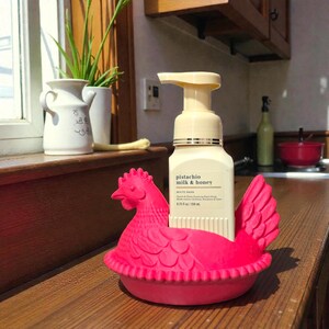 Puede incluir: Un jabonera en forma de gallina rosa brillante que sostiene una botella de jab&oacute;n de manos espumoso de leche de pistacho y miel. La jabonera est&aacute; hecha de un material gomoso y tiene un dise&ntilde;o detallado. La botella de jab&oacute;n es blanca con una bomba beige.