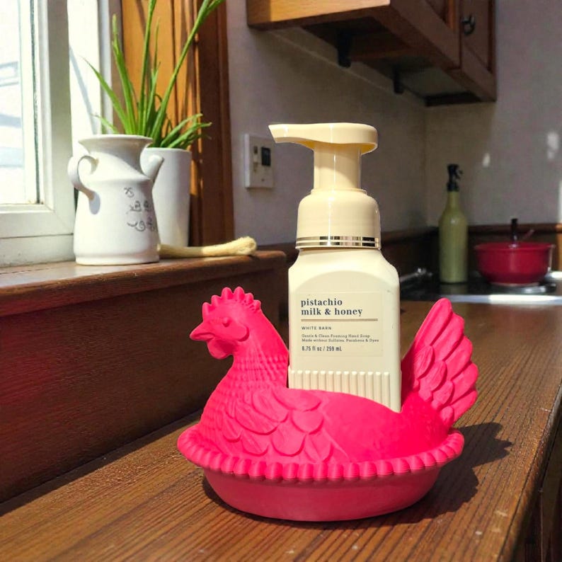 Puede incluir: Un jabonera en forma de gallina rosa brillante que sostiene una botella de jab&oacute;n de manos espumoso de leche de pistacho y miel. La jabonera est&aacute; hecha de un material gomoso y tiene un dise&ntilde;o detallado. La botella de jab&oacute;n es blanca con una bomba beige.