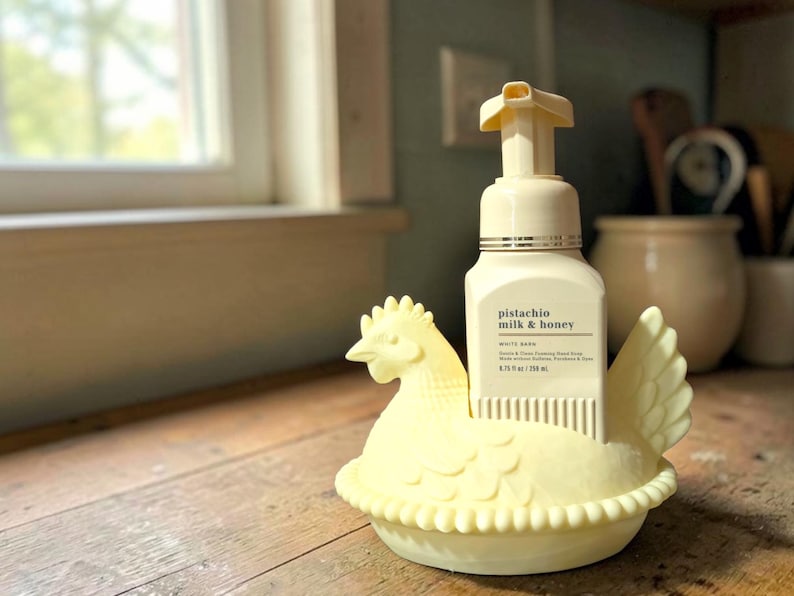 Puede incluir: Un jabonera en forma de gallina de color amarillo claro que sostiene una botella de jab&oacute;n de manos espumoso de leche y miel de pistacho. El dispensador de jab&oacute;n es blanco con una bomba amarilla. El plato est&aacute; sobre una superficie de madera.