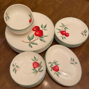 May include: A set of white ceramic dinnerware with a green and red apple design. The set includes a bowl, a plate, a saucer, and a larger plate.