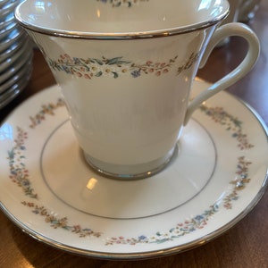 Può includere: Una tazza da tè e un piattino bianchi con un motivo floreale in tonalità di rosa, blu e verde. La tazza ha un bordo dorato e il piattino ha un bordo argentato.
