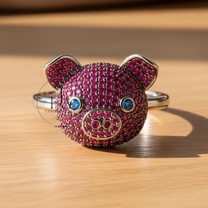 Puede incluir: Un anillo de plata con un diseño de cerdo. La cabeza del cerdo está cubierta de pequeñas piedras de color magenta, con piedras azules para los ojos. El cerdo tiene pequeñas orejas plateadas y una banda plateada.