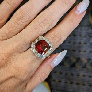 Puede incluir: Un anillo de plata con una gran piedra preciosa roja oscura de talla cuadrada, rodeada de piedras preciosas más pequeñas y claras. El anillo tiene piedras de acento rectangulares. El anillo se muestra en una mano con uñas largas y blancas.