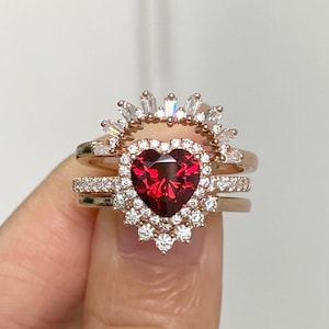 Puede incluir: Un anillo de oro rosa con una piedra roja en forma de corazón rodeada por un halo de piedras blancas más pequeñas. El anillo forma parte de un conjunto de tres piezas, con dos bandas adicionales que presentan piedras blancas más pequeñas.