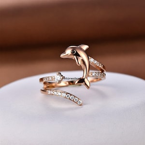 Può includere: Un anello in oro rosa con un design a forma di delfino e pietre scintillanti trasparenti. Il delfino salta fuori dall'acqua e l'anello si avvolge intorno al dito.