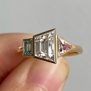 May include: A gold ring with a large, clear, rectangular-shaped gemstone, a smaller, light blue, rectangular-shaped gemstone, and a smaller, pink, triangular-shaped gemstone.