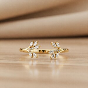 May include: A gold-colored ring with a leaf design. The ring features marquise-cut gemstones and small round gemstones. The ring is open at the front, with the leaf design extending from each side.