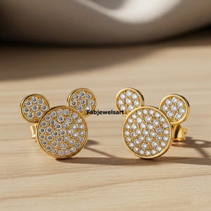 Puede incluir: Un par de pendientes de bot&oacute;n dorados con dise&ntilde;o de Mickey Mouse. Cada pendiente presenta un centro grande y redondo de circonita c&uacute;bica pavimentada con dos peque&ntilde;os detalles redondos de circonita c&uacute;bica pavimentada. Los pendientes est&aacute;n sobre una superficie de madera.