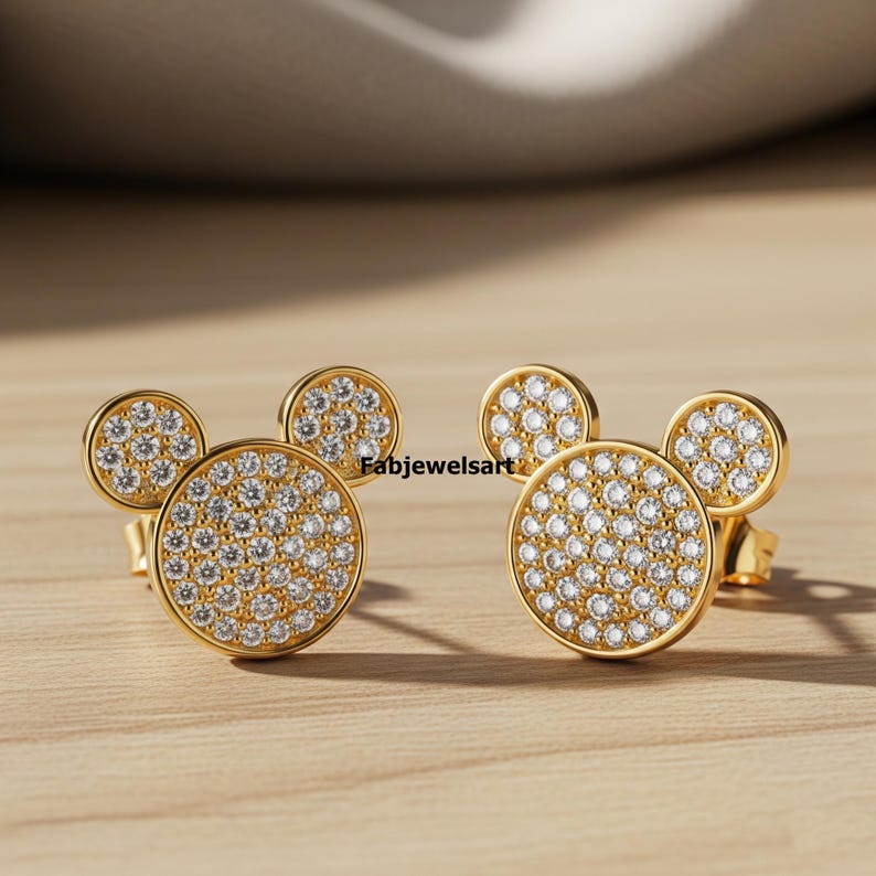 Puede incluir: Un par de pendientes de bot&oacute;n dorados con dise&ntilde;o de Mickey Mouse. Cada pendiente presenta un centro grande y redondo de circonita c&uacute;bica pavimentada con dos peque&ntilde;os detalles redondos de circonita c&uacute;bica pavimentada. Los pendientes est&aacute;n sobre una superficie de madera.
