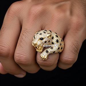 May include: A gold-coloured ring shaped like a jaguar, adorned with black and clear gemstones. The jaguar's head features an open mouth and visible teeth. The ring is worn on a finger.