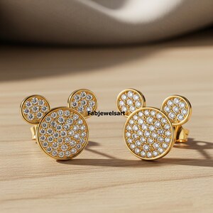 Puede incluir: Un par de pendientes de bot&oacute;n dorados con dise&ntilde;o de Mickey Mouse. Cada pendiente presenta un centro grande y redondo de circonitas c&uacute;bicas engastadas en pav&eacute; con dos peque&ntilde;os detalles redondos de circonitas c&uacute;bicas engastadas en pav&eacute;. Los pendientes est&aacute;n sobre una superficie de madera.