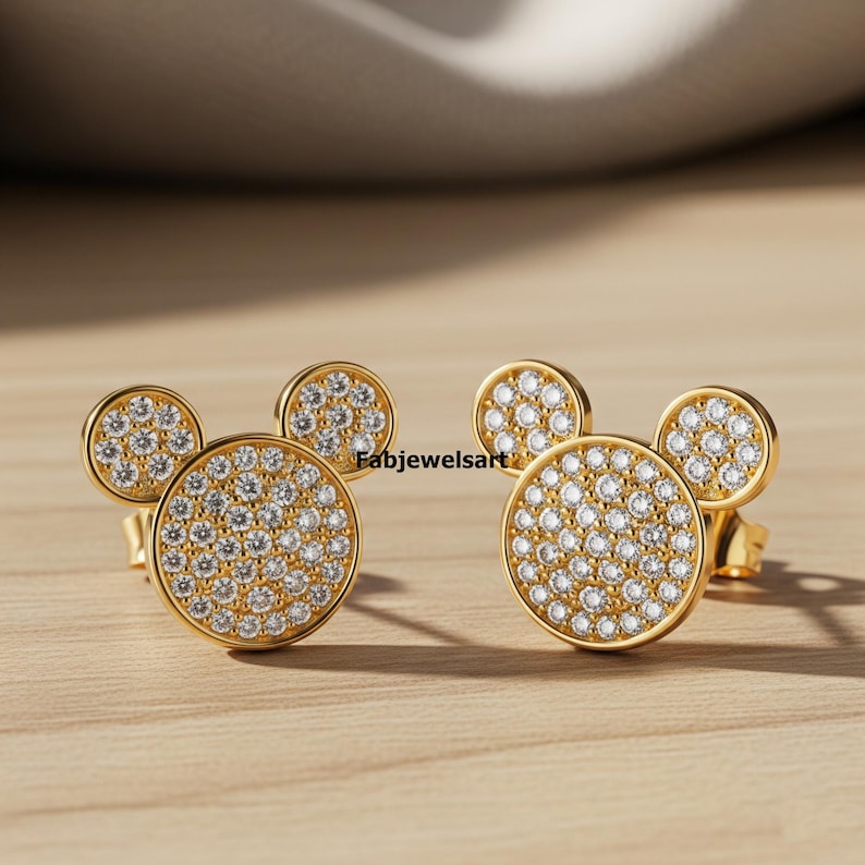 Puede incluir: Un par de pendientes de bot&oacute;n dorados con dise&ntilde;o de Mickey Mouse. Cada pendiente presenta un centro grande y redondo de circonitas c&uacute;bicas engastadas en pav&eacute; con dos peque&ntilde;os detalles redondos de circonitas c&uacute;bicas engastadas en pav&eacute;. Los pendientes est&aacute;n sobre una superficie de madera.