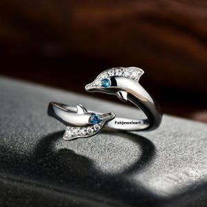 Può includere: Un anello d'argento con due delfini che si fronteggiano. I delfini sono adornati con piccole pietre trasparenti e ognuno ha una pietra blu per l'occhio. L'anello è aperto e regolabile.