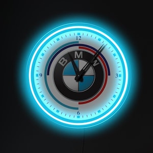 May include: A blue neon clock with a white face and the BMW logo in the center. The clock is hanging on a dark wall in a living room setting.