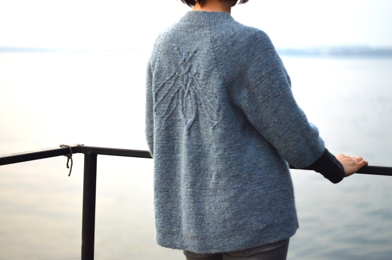以下が含まれることがあります： 背中が幾何学模様の青いニットセーター。セーターは、水面を見下ろすバルコニーに立っている人が着ています。