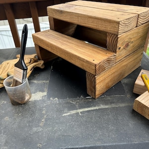 以下が含まれることがあります： 明るい茶色の木材で作られた2段の木製スツール。黒い柄の筆が、茶色の液体が入った透明なプラスチックカップに入っています。作業用手袋と、黄色の鉛筆が付いた2つの小さな木製ブロックも表示されています。