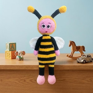 Pode incluir: Uma boneca de abelha em crochê com rosto rosa, corpo listrado de preto e amarelo, asas brancas e antenas longas com pontas amarelas. A boneca tem olhos brancos grandes com pupilas pretas e mãos e pés cor-de-rosa, feitos de fio texturizado.