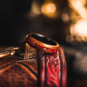 May include: A wooden ring with a red inlay. The ring is sitting on a brown leather box with a red and gold design.