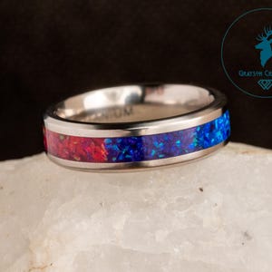 May include: A silver titanium ring with a red and blue opal inlay. The ring is on a white rock.