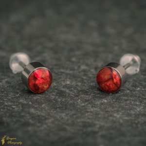 May include: A pair of small, round, red earrings with a silver backing. The earrings have a sparkly, iridescent finish.