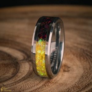 以下が含まれることがあります： 黒と黄色のインレイが付いた銀色のタングステンカーバイドリング。インレイは砕いたオパールで作られており、キラキラと輝いています。