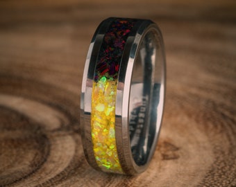 Könnte beinhalten: Ein silberner Wolframcarbid-Ring mit einer schwarzen und gelben Einlage. Die Einlage besteht aus zerkleinertem Opal und hat ein glitzerndes Aussehen.