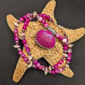 May include: A pink and white agate bead necklace with a pink seashell pendant. The necklace is draped over a starfish.