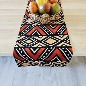 Peut inclure: Chemin de table coloré avec un motif géométrique rouge, noir, beige et blanc. Un panier en osier rempli d'oranges, d'une mangue, d'une poire, de kiwis et d'une banane est posé dessus. Le chemin de table est sur une table en bois clair.