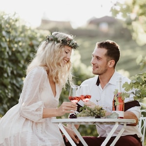 Pode incluir: Um casal desfrutando de uma degusta&ccedil;&atilde;o de vinhos ao ar livre. Est&atilde;o sentados a uma mesa branca com duas ta&ccedil;as de vinho ros&eacute;, uma garrafa de vinho e um cacho de uvas. A mulher usa uma coroa de flores e um vestido branco.