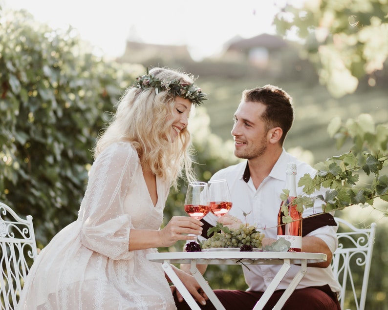 Pode incluir: Um casal desfrutando de uma degusta&ccedil;&atilde;o de vinhos ao ar livre. Est&atilde;o sentados a uma mesa branca com duas ta&ccedil;as de vinho ros&eacute;, uma garrafa de vinho e um cacho de uvas. A mulher usa uma coroa de flores e um vestido branco.