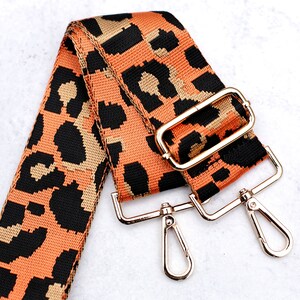 May include: A leopard print adjustable strap with gold-tone hardware. The strap is made of woven fabric and has a black, orange, and gold color scheme.