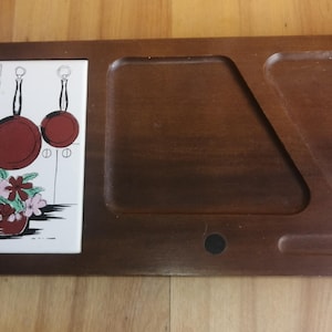 Peut inclure: Un plateau de service rectangulaire en bois avec un insert en carrelage décoratif. Le carreau présente une scène de cuisine avec une bouteille jaune, des casseroles rouges et une tasse bleue. Le plateau a une finition marron foncé.