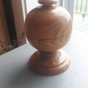 May include: A wooden box with a lid, made of light brown wood with a grain pattern. The box has a rounded shape and a smooth finish.