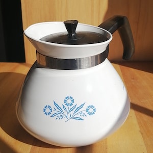 May include: A white teapot with a rounded body and a black lid featuring a central knob. The teapot has a black handle, a silver-colored metal band around the neck, and a blue floral pattern printed on the front of the white enamel surface.